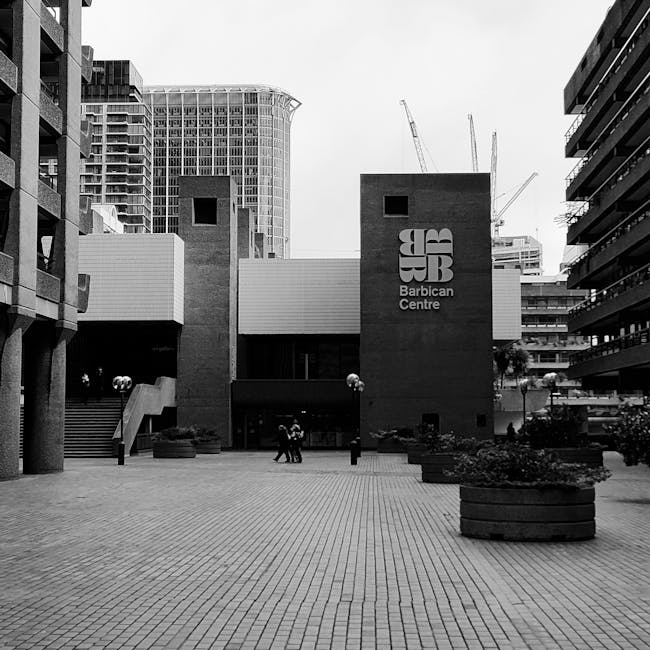 Black and white photograph of the exterior of the Barbican Centre on Aldersgate Street in the City of London, showing a modern architectural building with two prominent rectangular towers displaying the Barbican Centre logo. In front of the building, there is a paved courtyard with large round planters filled with plants, and several people walking or standing, some carrying items or engaging in conversation. The environment is urban, with taller buildings and construction cranes visible in the background, indicating ongoing development. The scene captures a moment of activity during a home relocation or moving process, with the area prepared for loading or unloading furniture and boxes, supported by the presence of [COMPANY_NAME], which specialises in removals and furniture transport in the Barbican area.