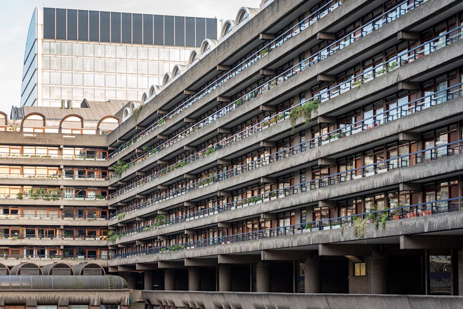 A multi-storey residential building with several rows of balconies featuring metal railings and glass panels, some with plants and outdoor furniture. The building's facade is composed of concrete with a weathered appearance, and large glass windows behind the balcony railings. In the background, a modern office building with a glass exterior is visible, contrasting with the older residential structure. The scene is lit with natural daylight, showing part of a loading zone at the building's ground level where Man and Van Barbican personnel may be engaged in house removals or furniture transport activities, although no moving process or personnel are visible in this specific image. The overall environment suggests an urban residential area suitable for home relocation or packing and moving logistics focused on apartment buildings.