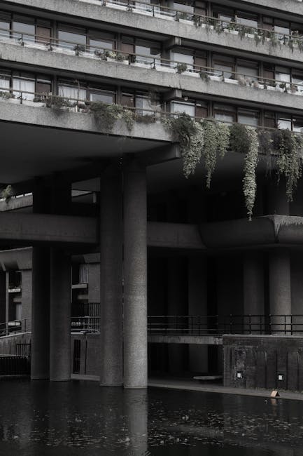 A black-and-white photograph of a modern, multi-storey residential building with protruding balconies and horizontal railings, featuring some greenery cascading from balcony planters. The building's structure includes large concrete pillars supporting the upper levels and a dark underside that creates shadows. In the foreground, a reflection of the lower part of the building and the water surface is visible, indicating a nearby water feature or canal. The image captures a section of the exterior environment, emphasizing the architectural design and urban setting. This scene relates to house removals, as Man and Van Barbican provides professional relocation services that include handling the transport of furniture and belongings through such complex building environments, ensuring safe and efficient home relocation or furniture transport for residents of the Golden Lane Estate in the Barbican area.