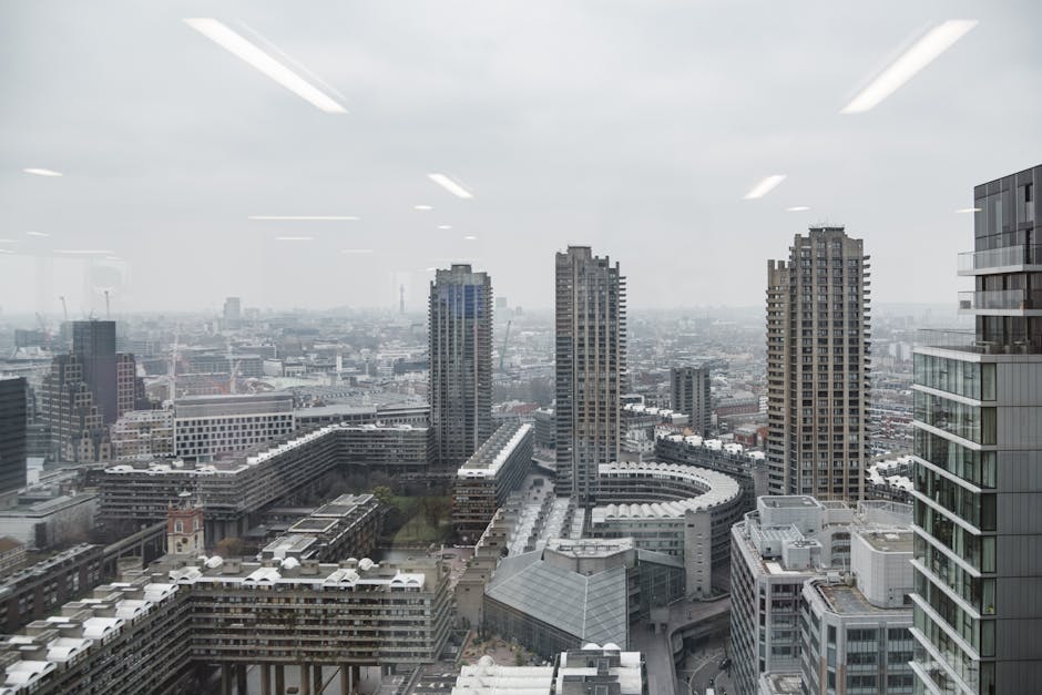 A cityscape view of high-rise buildings in an urban area with modern architectural designs, seen from inside a property through a large window. The photograph shows several tall residential and commercial towers, some with glass facades reflecting the overcast sky, and shorter buildings near the foreground featuring flat roofs and balconies. Light reflections of ceiling-mounted fluorescent lights are visible on the window's interior surface. The scene captures the metropolitan environment typical of a London neighbourhood such as the Golden Lane Estate, with the distant skyline blending into a grey, cloudy atmosphere. This view provides context for local house removals or furniture transport, with the window serving as a point of observation before the logistics of home relocation or loading furniture into a vehicle by [COMPANY_NAME], such as a van for property moving and packing and moving processes.