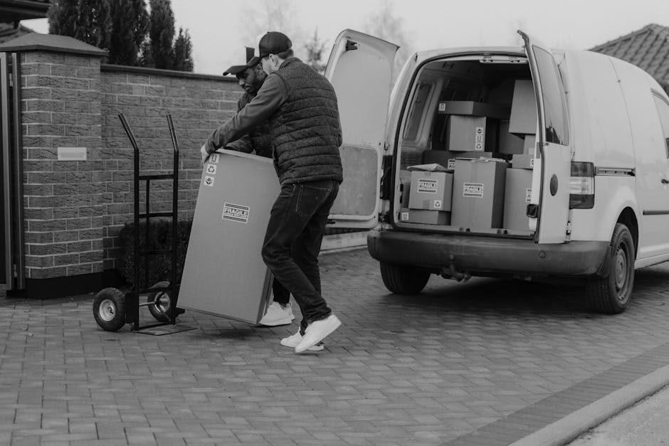 A young man with long, wavy hair and a black headband is sitting in the driver's seat of a white moving van labeled 'Moving Company Local & Long Distance,' parked outside a residential building in an 