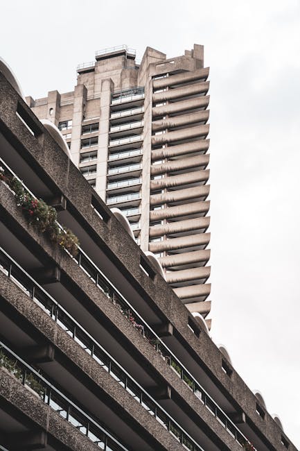 A low-angle view of a high-rise residential building with multiple balconies, some decorated with plants and outdoor furniture, set against an overcast sky. In the foreground, part of a concrete balcony with a metal railing is visible, showing potted plants with flowers. The image captures a section of a multi-storey parking structure with curved concrete edges and metal railings on the lower levels. Inside a property, a team from Man and Van Barbican is engaged in furniture transport as part of a home relocation or packing and moving process, with a large cardboard box and protective wrapping visible in the background near the entrance. The loading process appears to involve lifting and carrying items, supported by the presence of moving blankets and straps. The scene reflects the logistical aspects of professional removals within an urban environment, highlighting the importance of careful handling and efficient transportation during house removals, particularly when facing scheduling delays or complex building access issues.
