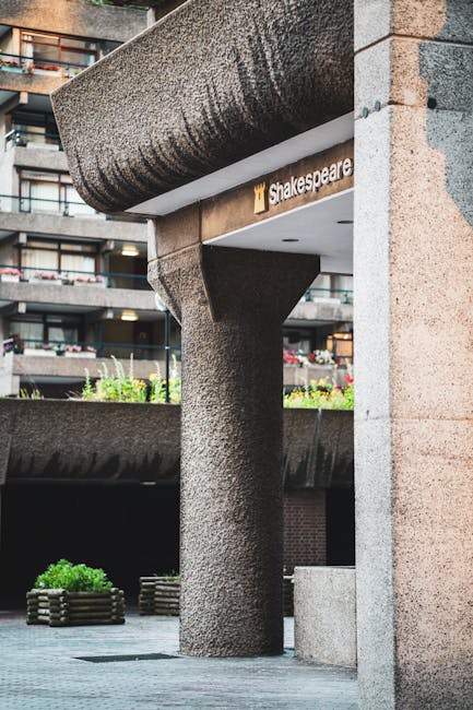 An exterior view of a building entrance showing a textured concrete column supporting an overhanging canopy with a rounded edge. The canopy has a reflective under-surface, and a sign attached beneath it displays the name 'Shakespeare.' In the background, the building features multiple balconies with glass railings and potted plants, indicating urban residential architecture typical of a house removal or relocation environment. Near the base of the column, there are wooden planters with green foliage, and the pavement around the area is clean and well-maintained. This setting reflects the site where furniture or packing materials might be prepared for loading into a removal van, captured during daylight hours with good natural lighting. The scene is associated with moving logistics, where coordination of loading furniture or boxes could take place outside this location, potentially involving companies like Man and Van Barbican, specialists in removals in the area.