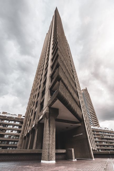 A low-angle view of a high-rise residential building with multiple balconies, some decorated with plants and outdoor furniture, set against an overcast sky. In the foreground, part of a concrete balcony with a metal railing is visible, showing potted plants with flowers. The image captures a section of a multi-storey parking structure with curved concrete edges and metal railings on the lower levels. Inside a property, a team from Man and Van Barbican is engaged in furniture transport as part of a home relocation or packing and moving process, with a large cardboard box and protective wrapping visible in the background near the entrance. The loading process appears to involve lifting and carrying items, supported by the presence of moving blankets and straps. The scene reflects the logistical aspects of professional removals within an urban environment, highlighting the importance of careful handling and efficient transportation during house removals, particularly when facing scheduling delays or complex building access issues.