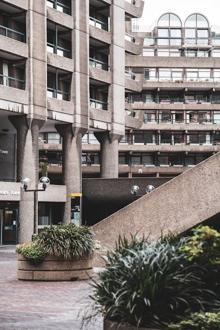 The image shows a modern urban building complex with a concrete exterior, featuring multiple structures of varying heights, including a tall, rectangular tower with small, evenly spaced windows. In th