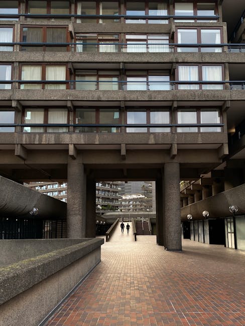 A multi-storey residential building with concrete balconies lined with blue metal railings, each decorated with potted plants and flowers. The building has a weathered appearance with some visible moss and dirt on the concrete surfaces. Large glass windows can be seen behind the balconies, reflecting the surrounding urban environment. In the background, there are modern office buildings with reflective glass facades. The scene is captured during daylight with clear weather. This image depicts a typical urban residential block in the City of London area, relevant to house removals and furniture transport services provided by Man and Van Barbican, highlighting the need for careful planning during home relocation in such environments.