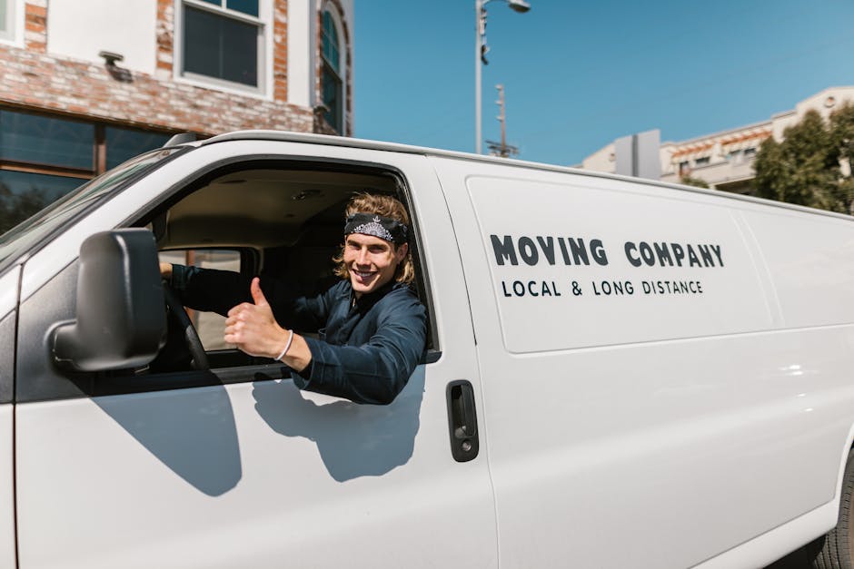 Inside a white moving van labeled with the company name 'Moving Company' and the description 'Local & Long Distance,' two men are visible preparing for a home relocation. The man sitting in the driver