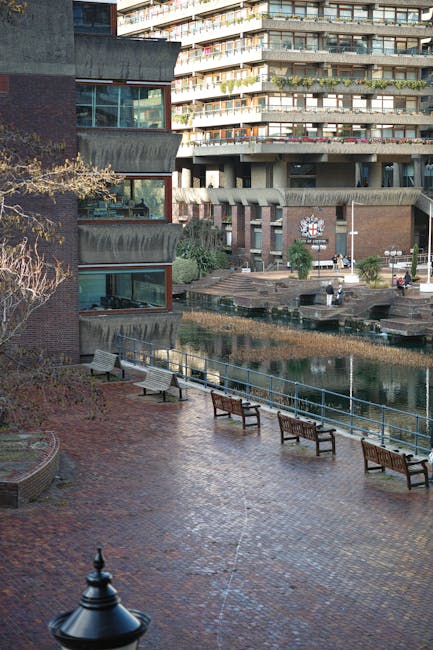 The image depicts a riverside area adjacent to modern multi-storey residential buildings, with several wooden and metal benches arranged along the paved walkway overlooking the water. In the foreground, there is a black lamp post, and leafless trees with some greenery are visible on the left side. The background features a concrete and glass apartment complex with balconies facing the water, and a smaller brick building with a clock on its façade. The scene is well-lit with natural daylight, and there are no people visible. This setting may be part of a housing estate or residential complex where home relocation or furniture transport services, such as those offered by Man and Van Barbican, might take place as part of moving logistics.