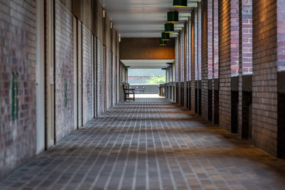 A long, covered outdoor corridor with brick walls on both sides and a series of evenly spaced green rectangular light fixtures hanging from the ceiling. The floor is paved with reddish-brown bricks arranged in a pattern, and the corridor is illuminated by natural light coming from an open outdoor area at the far end, where a wooden bench and a small table are placed near a railing overlooking a bright, green outdoor space. In the foreground, there are some cardboard boxes and loose packing materials arranged along the corridor, suggesting ongoing house relocation or furniture transport preparations. The scene appears to be part of a property exit or loading zone, typical in moving or removals services such as those provided by Man and Van Barbican, as indicated by the context of the service category and webpage URL.