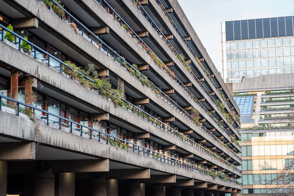 The image depicts a riverside area adjacent to modern multi-storey residential buildings, with several wooden and metal benches arranged along the paved walkway overlooking the water. In the foreground, there is a black lamp post, and leafless trees with some greenery are visible on the left side. The background features a concrete and glass apartment complex with balconies facing the water, and a smaller brick building with a clock on its façade. The scene is well-lit with natural daylight, and there are no people visible. This setting may be part of a housing estate or residential complex where home relocation or furniture transport services, such as those offered by Man and Van Barbican, might take place as part of moving logistics.