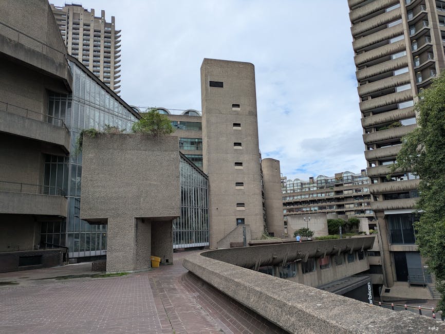 The image shows a tall modern high-rise building in the background, with a low-rise, curved residential complex in the foreground featuring multiple levels with balconies and dark exterior walls. Seve