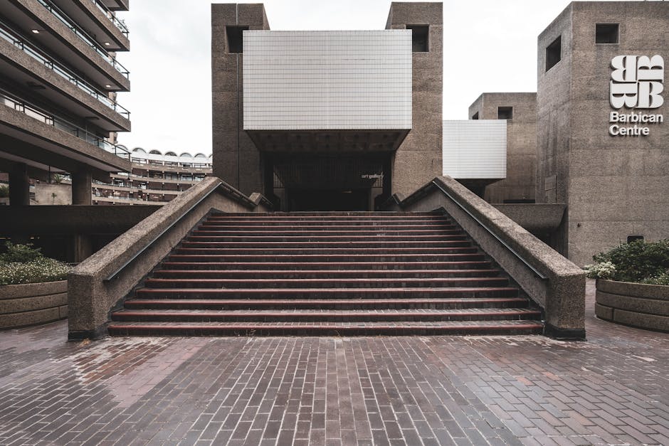 A wide set of outdoor concrete stairs with red-tinted steps leading up to the entrance of the Barbican Centre, situated in a modern urban complex. The stairs are flanked by concrete railings on both sides, with large concrete planters filled with greenery at the bottom. The surrounding environment includes adjacent residential or office buildings with multiple balconies and large windows, characteristic of the Barbican estate. The area appears slightly wet, indicating recent rain, with overcast lighting giving a neutral tone to the scene. The entrance to the building features a dark opening beneath a rectangular overhang, and prominent signage displaying the Barbican Centre logo is visible on the right. This setting is typical for a home relocation or furniture transport process, where access issues might be considered, and [COMPANY_NAME] may assist with removals or moving logistics, particularly in complex urban environments such as this one.