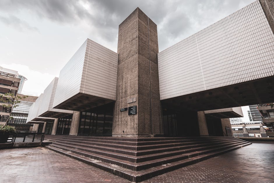 The exterior of a modern commercial building with a central concrete column and white tiled facades extending from the sides. The structure features multiple levels with a staircase leading up to the entryway. The entrance area is paved with dark bricks, and there are glass doors at the base of the building. The building's overhanging sections are supported by concrete columns, creating a sheltered loading zone underneath. Surrounding the building are other city structures and parked vehicles, indicating an urban environment. The scene is captured during daylight, with overcast skies casting diffused light. This image exemplifies the type of location where professional removals services, such as those provided by Man and Van Barbican, are often required for efficient home relocation or furniture transport, especially when access challenges are present.